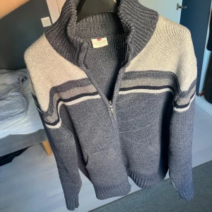Grå kofta från Woolpark i ull - Säljer en gråmelerad kofta från Woolpark i 100% ull. Koftan har dragkedja framtill, ståkrage och ribbade muddar. Snygga ljusgrå ränder över axlar och bröst ger en sportig vibe. Perfekt för kyliga dagar och enkel att matcha med jeans.
