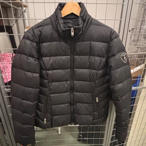 Svart Jacka från Rockandblue - Svart jacka från Rockandblue med quiltad design och hög krage. Jackan har två sidofickor med dragkedja och en snygg patch på vänster ärm. Perfekt för kalla dagar och har en modern, sportig look.