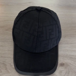 Svart keps från Fendi med logomönster - Snygg svart keps från Fendi med diskret FF-logomönster över hela kepsen. Justerbar rem baktill i svart skinn med Fendi-tryck. Kepsen är tillverkad i bomull och har klassisk böjd skärm. Perfekt accessoar för en trendig streetstyle-look.
