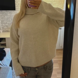 Beige polotröja  - Mysig beige stickad polotröja med ribbad struktur och lång ärm. Tröjan har en loose passform och hög krage som ger en chill och trendig vibe. Perfekt för lager-på-lager under kyliga dagar.
