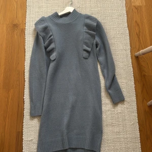 Blå stickad klänning med volang Lindex - Mysig blå stickad klänning från Lindex med långa ärmar och volangdetaljer på axlarna. Ribbstickad i hals, ärmslut och nederkant. Använd en gång 🩷