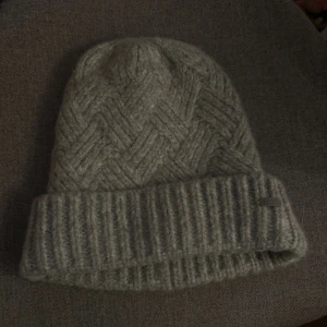 Grå stickad mössa från KAVAT - Säljer en snygg grå stickad mössa från KAVAT med bred uppvikt kant och ett stilrent fiskbensmönster. Mössan är gjord i mjuk ullblandning och har en liten metallbricka med logga framtill. Perfekt för kalla dagar och passar till allt. Är inte andvänd!
