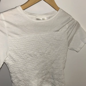 Vit smockad topp från Gina Tricot - Vit topp från Gina Tricot med smockad midja och korta ärmar. Den är i bra skick och har inga fläckar eller hål och är i storlek 158/164 men den är stretchig och formar sig efter ens kropp. 