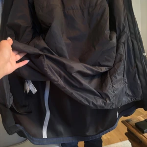 Svart parkajacka med dragkedja - Svart parkajacka med hög krage och dragkedja framtill, kompletterad med tryckknappar. Jackan är längre i modellen och har en stilren look. Tillverkad i ett slätt syntetmaterial som känns tåligt och passar perfekt för kyligare och varmare dagar. Jackan är både sommar och vinter jacka, man kan välja själv om man vill behålla den tunn eller tjock då man kan knäppa bort ett lager❤️