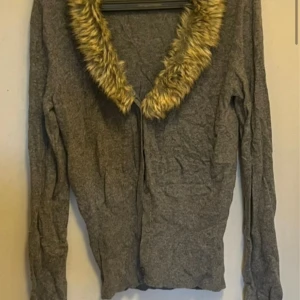 Grå kofta med fuskpäls från Amisu - Säljer en grå kofta från Amisu i storlek XL med långärmad design och en snygg krage i beige fuskpäls. Perfekt för att ge din outfit en extra touch. Koftan är mjuk och skön, passar bra till kyligare dagar.