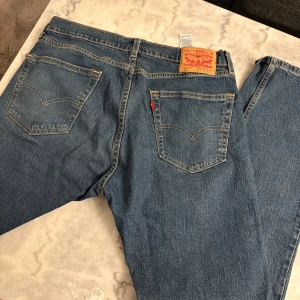 Levi's 501 blå jeans straight fit - Klassiska blå Levi's 501 jeans med raka ben och femficksmodell. Jeansen har den ikoniska läderpatchen bak, synliga nitar och gul kontrastsöm. Tillverkade i robust bomullsdenim med normal passform och knappgylf. Perfekta för en avslappnad och tidlös look. Knappt använd!