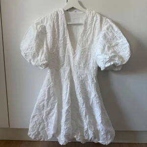 Vit kort klänning med puffärmar - Superfin vit kort klänning med stora puffärmar och v-ringning. Klänningen har broderade blommor över hela tyget och en lätt voluminös kjol. Perfekt att ha som studentklänning eller i sommar!🌸