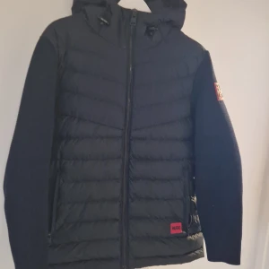 Svart pufferjacka från HUGO, storlek L - Snygg svart pufferjacka från HUGO med huva och dragkedja. Jackan har quiltad front i syntetmaterial och stickade ärmar med röd HUGO-logga på vänster arm. Perfekt för höst och vinter, riktigt modern look och skön passform.