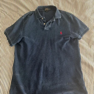 Marinblå pikétröja Polo Ralph Lauren M - Säljer en marinblå pikétröja från Polo Ralph Lauren i slim fit. Klassisk krage, kort ärm och två knappar framtill. Ikoniska röda loggan broderad på bröstet. Perfekt för dig som gillar stilren och sportig look.