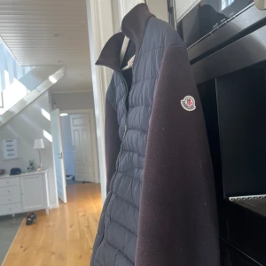 Svart Moncler pufferjacka med stickade ärmar - Snygg svart pufferjacka från Moncler med quiltad front och stickade ärmar. Jackan har hög krage, dragkedja framtill och Moncler-logga på ärmen. Perfekt för dig som vill ha en stilren och sportig look med premiumkänsla.Pris inte ristat i sten så tveka inte skicka in frågor och funderingar samt prisförslag