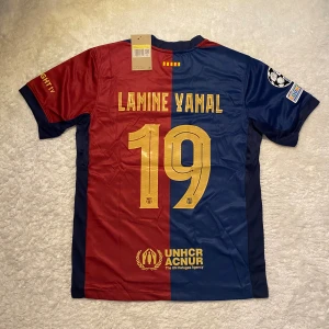FC Barcelona Lamine Yamal fotbollströja  - Säljer en officiell FC Barcelona fotbollströja från Nike med Lamine Yamal och nummer 19 på ryggen. Tröjan är kortärmad, i röd och mörkblå färg, med klubbmärke, Champions League-patch och sponsorer som Spotify och UNHCR. Tillverkad i Dri-FIT-material för bästa komfort.