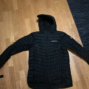 Svart pufferjacka Peak Performance - Svart pufferjacka från Peak Performance med huva och dragkedja framtill. Jackan har quiltad design och logga på bröstet samt ärmen. Perfekt för kyliga dagar och enkel att matcha med allt.