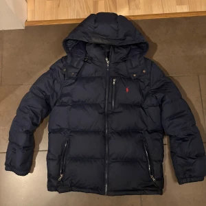 Mörkblå Polo Ralph Lauren - Säljer en mörkblå pufferjacka från Polo Ralph Lauren med huva och flera dragkedjefickor. Jackan har det klassiska röda Polo-loggan broderad på bröstet och är perfekt för kalla dagar. Stängs med dragkedja och har en schysst, vadderad look.