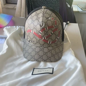 Gucci keps kingsnake - Säljer en limited edition kingsnake keps från Gucci som ej SÄLJS LÄNGRE med klassiskt GG-mönster i beige och brunt, prydd med en röd orm framtill. Baksidan är i svart mesh och justerbar med grön-röd randig rem. Perfekt för dig som vill sticka ut med en lyxig accessoar. 