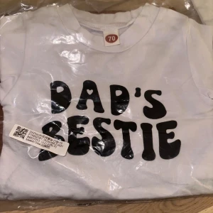 Vit t-shirt med text Dad's Bestie - Vit t-shirt i bomull med svart tryck 'Dad's Bestie' på bröstet. Klassisk rund halsringning och korta ärmar. Perfekt för en cool och avslappnad look till de minsta.