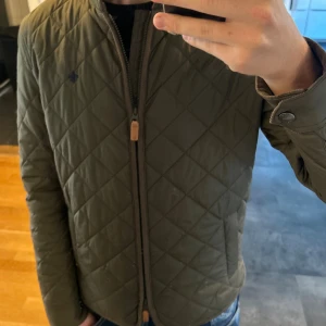 Olivgrön quiltad jacka från Morris - Säljer en olivgrön quiltad jacka från Morris i storlek Small. Jackan har ett stilrent rutmönster, dragkedja framtill och snygga detaljer vid ärmsluten. Perfekt för dig som vill ha en klassisk look med modern touch.