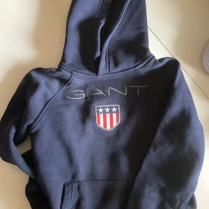 Marinblå hoodie från GANT med logga - Snygg marinblå hoodie från GANT med broderad logga och sköld på bröstet. Tröjan har huva och en klassisk känguruficka framtill. Perfekt för dig som gillar stilrena och sportiga plagg med preppy känsla.