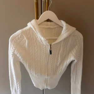 Vit stickad hoodie med dragkedja - Supermysig vit stickad hoodie med dragkedja framtill och kabelstickat mönster. Tröjan har ribbade muddar, huva och lång ärm. Perfekt för lager-på-lager och chill dagar.