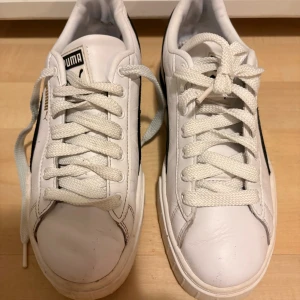 Puma vita sneakers med svart detalj - Säljer ett par snygga Puma sneakers i vitt skinn med svarta detaljer längs sidan och guldigt Puma-logo. Skorna har platt sula, rund tå och klassisk snörning. Perfekt för dig som gillar stilrena och sportiga sneakers med chunky look. Innersula saknas