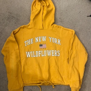Gul hoodie från Gina Tricot XS - Gul hoodie från Gina Tricot med trycket 'The New York Wildflowers' och amerikansk flagga framtill. Tröjan har huva, långa ärmar och snörning i nederkanten. Perfekt för dig som gillar streetstyle och vill sticka ut med en färgglad look.