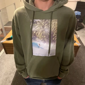 Olivgrön hoodie med surftryck från Bvaldi - Olivgrön hoodie från Bvaldi med stor surf-inspirerad print framtill. Tröjan har huva med snörning, känguruficka och långa ärmar. Perfekt för dig som gillar chill och streetwear vibes.
