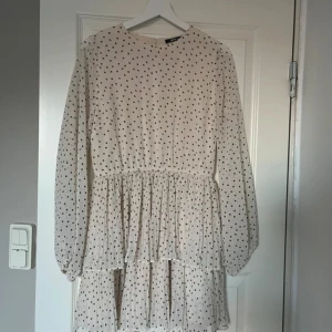 Prickig kortklänning från Gina Tricot - Supersöt kortklänning från Gina Tricot i ljus färg med svarta prickar. Klänningen har långärmade ärmar, rund hals och volangdetaljer i kjolen. Perfekt för dig som gillar en lekfull och trendig stil. Tillverkad i ett lätt och luftigt material. Något sksvd i tyget, se sista bilden