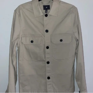 Beige overshirt från H&M XS - Stilren beige overshirt från H&M i storlek XS. Jackan har två stora bröstfickor med knappar, klassisk krage och svarta knappar längs hela framsidan. Perfekt lager-på-lager-plagg med lång ärm och rak passform.
