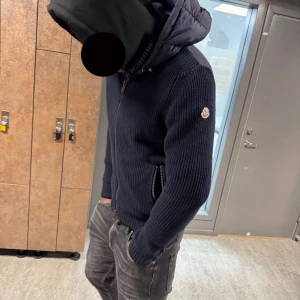 Mörkblå Moncler cardigan äkta - Säljer en mörkblå stickad cardigan jacka från Moncler med huva och dragkedja framtill. Jackan har ribbad struktur, Moncler-logga på ärmen och två fickor med dragkedja. Perfekt för dig som vill ha en snygg och bekväm jacka med street-vibe.