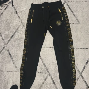 Svarta mjukisbyxor med gulddetaljer - Svarta mjukisbyxor med guldfärgade dragkedjor och snörning i midjan. Sidorna har glittriga guldfärgade mönster och ett märke på låret. Byxorna har ribbade muddar vid bensluten och en slim passform. Perfekta för en trendig och bekväm stil.