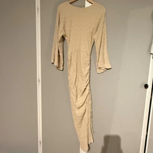 Beige långklänning från NA-KD - Superfin beige långklänning från NA-KD med strukturerat tyg, vida långa ärmar och snygg slits med knyt i sidan. Klänningen har en figurnära passform med rynkade detaljer längs midjan och höften. Använd endast en gång, nypris 850kr