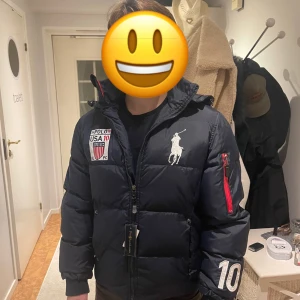 Mörk blå pufferjacka Polo Ralph Lauren - Mörk blå  pufferjacka från Polo Ralph Lauren med broderade loggor och USA-märke på bröstet. Jackan har dragkedja framtill, huva och detaljer som broderad siffra på ärmen samt röd dragkedja på ena ärmen. Perfekt för kalla dagar.