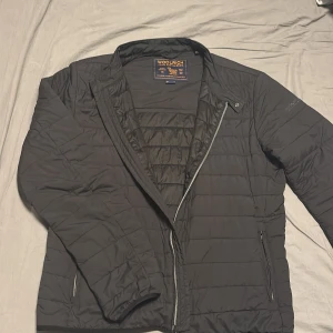 Svart quiltad dunjacka från Woolrich - Snygg svart quiltad dunjacka från Woolrich med dragkedja och diskret logga på ärmen. Jackan har en klassisk passform, två fickor med dragkedja och är perfekt för kyliga dagar. Lätt och smidig att bära, passar till de flesta outfits.