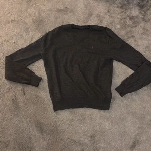 Mörkgrå stickad tröja Polo Ralph Lauren - Mörkgrå stickad tröja från Polo Ralph Lauren med v-ringning och diskret röd logga på bröstet. Tröjan har långa ärmar och ribbade muddar. Perfekt för lager-på-lager och chill stil. Notera att tröjan har krympt och därmed passar bäst som XS och möjligen S