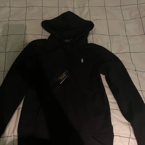 Svart hoodie från Polo Ralph Lauren - Svart hoodie från Polo Ralph Lauren med klassisk broderad logga på bröstet. Tröjan har huva med snörning och dragkedja framtill. Tillverkad i mjuk bomull, perfekt för en chill och stilren look.