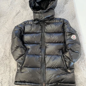 Svart Moncler dunjacka med huva Barn - Snygg svart dunjacka från Moncler med glansig finish, huva och dragkedja framtill. Jackan har Moncler-logga på ärmen, två fickor med dragkedja och quiltad design. Perfekt för kalla dagar när du vill hålla stilen.