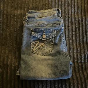Ljusblå bootcut jeans med broderad ficka - Säljer ett par ljusblå jeans med bootcut passform och snygg broderad bakficka med lock och knapp. Jeansen har klassisk femficksdesign och är tillverkade i mjukt denimtyg. Perfekta för dig som gillar en avslappnad men ändå trendig look.