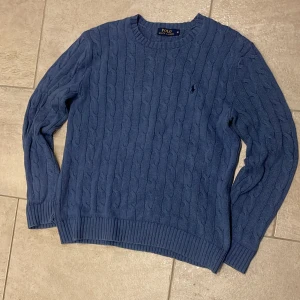 Blå kabelstickad tröja Polo Ralph Lauren - Kabelstickad blå tröja från Polo Ralph Lauren med klassisk rund halsringning och broderad logga på bröstet. Tröjan har ribbade muddar och är tillverkad i mjuk bomull. Perfekt för lager-på-lager och snygg till jeans.