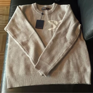 Beige stickad tröja Louis Vuitton S - Beige stickad tröja från Louis Vuitton med rund halsringning och ribbade muddar. Tröjan har en stor, vit LV-logga på bröstet och är långärmad. Perfekt för dig som gillar exklusiva och stilrena plagg med lyxig känsla.