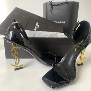 Svarta Saint Laurent sandaler med YSL-klack - Säljer ett par svarta sandaler från Saint Laurent i lackat skinn med ikonisk guldig YSL-klack. Skorna har öppen tå, smal rem över foten och runt vristen samt fyrkantig tå. Perfekta för dig som vill sticka ut med lyxig design och cool detalj.