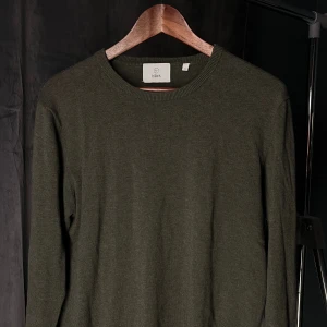 Olivgrön stickad tröja från Bläck - En stilren olivgrön stickad tröja från Bläck med rund halsringning och långa ärmar. Tröjan har en klassisk passform och är perfekt för lager-på-lager. Enkel design utan mönster eller detaljer, vilket gör den lätt att matcha med allt.