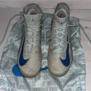 Nike Air Zoom fotbollsskor grå/blå - Säljer ett par Nike Air Zoom fotbollsskor i grått med blå detaljer och stor blå Swoosh på sidan. Skorna har snörning, högre skaft och en platt sula med mönstrad undersida för extra grepp. Materialet är syntet och mesh för lätthet och ventilation. Medföljer en matchande Nike-påse.