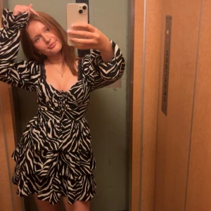 Zebramönstrad kortklänning med volanger - Säljer en snygg kort klänning med zebramönster i svart och vitt. Klänningen har långa puffärmar, v-ringad hals och knytning framtill. Volanger i kjolen ger extra flärd och en cool look. Perfekt för dig som vill sticka ut med ett djuriskt mönster.