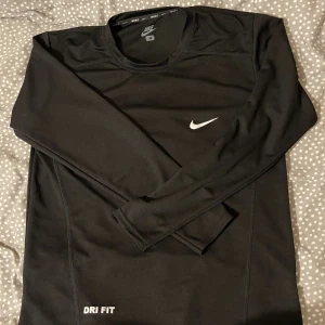 Svart långärmad Nike Dri-Fit tröja - Svart långärmad tröja från Nike med Dri-Fit-teknologi. Tröjan har rund halsringning, diskret Nike-logga på bröstet och är tillverkad i ett lätt och funktionellt syntetmaterial som andas. Perfekt för träning eller sportiga dagar.