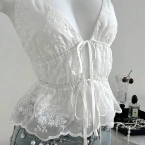 Vit blommig spets-linnetopp med snörning - Superfin vit linnetopp med tunna axelband och djup v-ringning. Toppen har broderade blommor i spets, snörning framtill och en lätt volangkant nedtill. Perfekt för dig som gillar romantisk och feminin stil. Snygg att matcha med jeans eller kjol.