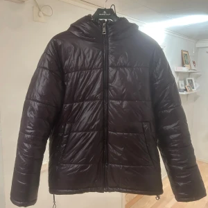 Svart pufferjacka med huva - Säljer nu min svarta pufferjacka. Jackan har huva, dragkedja framtill och två stora fickor. Den har en glansig finish. Perfekt för kalla dagar när du vill hålla dig varm men ändå ha bra stil. Pris kan sänkas vid snabb affär! 