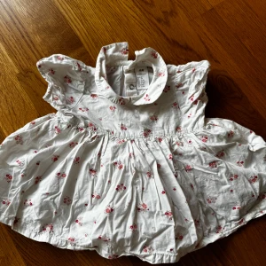 Vit blommig kort klänning från H&M - Supersöt vit kort klänning från H&M med små rosa blommor och volangkrage. Klänningen har korta ärmar och praktiska tryckknappar både bak och i grenen. Perfekt för varma dagar och enkel att ta på och av.