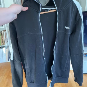 Svart hoodie från Hugo Boss med dragkedja - Svart hoodie från Hugo Boss med vit logga på bröstet och vita detaljer längs ärmarna. Tröjan har huva, hel dragkedja framtill och två fickor.  Strl M men sitter som S 