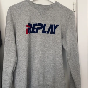Grå sweatshirt från Replay - Snygg grå sweatshirt från Replay med blå och röd logga framtill och liten logga på ärmen. Tröjan har rund halsringning och långa ärmar. Perfekt för en avslappnad och stilren look. Aldrig använd! Priset går att diskutera vid en snabb affär! 