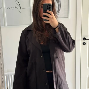 Svart parkajacka med snörning - Svart parkajacka med långa ärmar, knappar framtill och snörning i midjan för en snygg passform. Jackan har klassisk krage och är tillverkad i ett mjukt tyg som känns bekvämt. Perfekt för dig som gillar en stilren och enkel look.