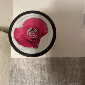 The Body Shop body yoghurt med rosdoft - En body yoghurt från The Body Shop med rosdoft, levereras i en rund burk med svart lock och en stor rosa ros på locket. Krämen är ljusrosa och har en lätt, krämig konsistens som känns återfuktande och fräsch på huden. Den är med doften birtish rosé och är aldrig använd